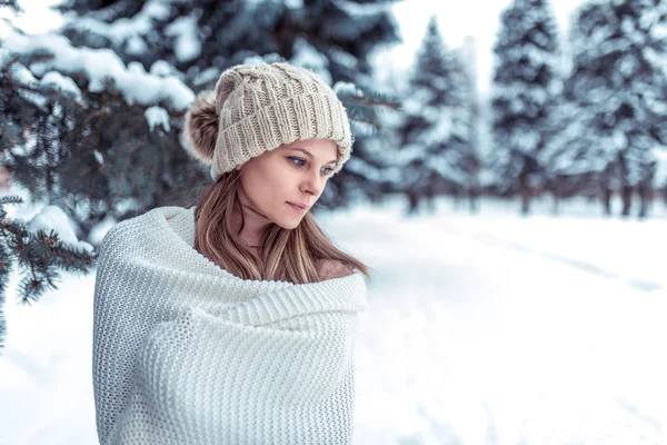 Krásná dívka v bílém plaid zabalený porostů v zimě na čerstvém vzduchu v zasněženém lese, na pozadí zelené vánoční stromky. Volné místo pro text. Béžový klobouk rád odpočívá. — Stock fotografie