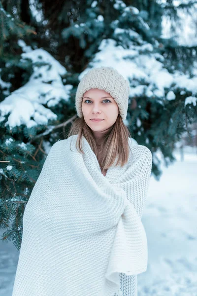 Een mooi meisje in een witte plaid verpakt staat in de winter in de frisse lucht in een besneeuwde forest, tegen een achtergrond van groene kerstbomen. In een beige hoed rust gelukkig. — Stockfoto