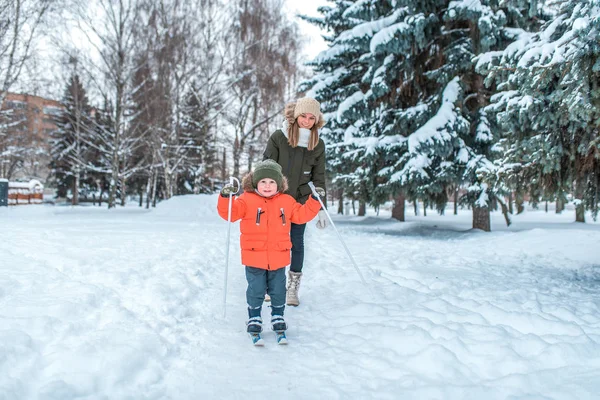 Menino de 3 a 5 anos, esquiando no inverno em uma floresta nevada. Uma jovem mãe entra pelas traseiras. Esqui ativo, natureza em um dia de inverno . — Fotografia de Stock
