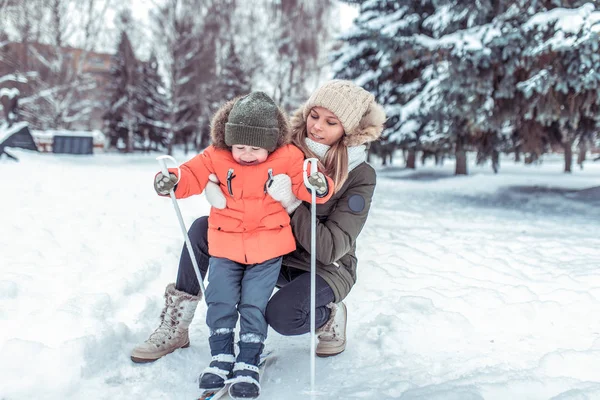 Seorang anak kecil berusia 3-5 tahun, naik ski untuk pertama kalinya, di musim dingin di hutan bersalju. Seorang ibu muda menggendong seorang anak di belakang. Berski aktif, alam pada hari musim dingin. Selamat tersenyum . — Stok Foto