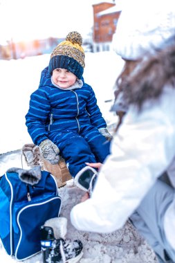 A little boy is sitting on a bench in a warm jumpsuit and a hat. Mom puts the skates on her child, changes her shoes. Tying shoelaces. In winter in the city on a public skating rink.
