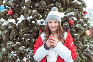 Genç bir kız ile bir sıcak şapka şehir kış orman içinde yeşil ağaç arka plan kırmızı ceketli. Bir fincan kahve ya da çay elinde tutar ve soğuk havalarda ısıtır. Mutlu gülümsemeler.