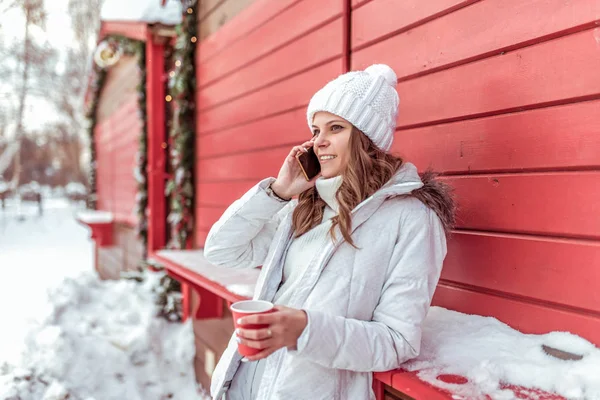 Piękna i młoda dziewczyna w białym kapeluszu i ciepłą kurtkę, w zimie po mieście w pobliżu czerwone ściany. W dłoniach trzyma kubek gorącej herbaty lub kawy. Dzwoniąc na telefon, szczęśliwy uśmiechający się. — Zdjęcie stockowe