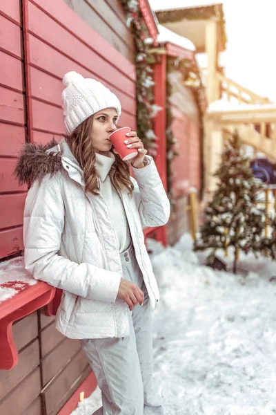 Beautiful and young girl in a white hat and warm jacket, in the winter in the city near the red wall. In his hands holding a cup of hot tea or coffee. Drinking from a mug. — Zdjęcie stockowe