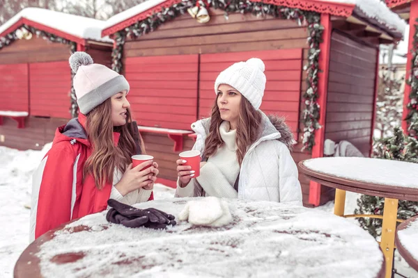 Dua pacar di musim dingin di udara segar dengan jaket hangat dan topi. Mereka beristirahat di dekat kafe, memegang cangkir dengan kopi panas atau teh di tangan mereka. Berkomunikasi berbicara dan pemanasan setelah berjalan . — Stok Foto