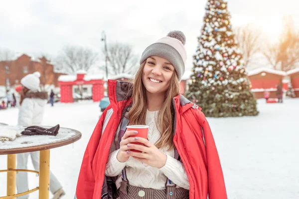 Młoda dziewczyna w czerwonej kurtce, ciepła czapka, w mieście zimą na tle zielonej choinki. W jego ręce, trzymając kubek gorącej herbaty lub kawy. Szczęśliwy, uśmiechnięty, spoczywającej na wakacje. — Zdjęcie stockowe