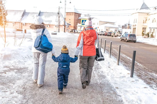 Две подружки мама сопровождать маленького мальчика ребенка 3-5 лет. Родители-женщины держатся за руки, зимой едут в город по заснеженной дороге рядом с машинами, в сопровождении дома, вид сзади . — стоковое фото