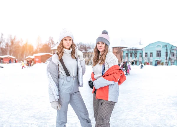 Dua gadis pacar musim dingin di kota dalam udara segar. Berpose dalam jaket dan topi, tersenyum. Dalam sweater hangat dan topi. Selamat tersenyum. Istirahat di kota di arena . — Stok Foto