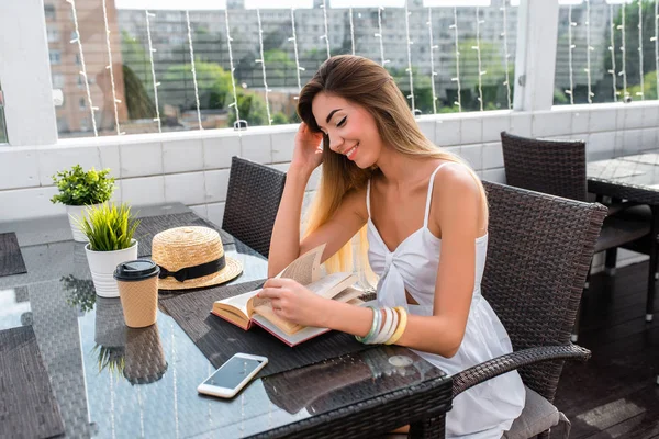 Menina bonita sentada no café da cidade no verão. Lê um livro de papel, ao lado de uma xícara de café ou chá e um smartphone. Sorrisos felizes. Senhora de negócios em ordem à espera. Pequeno-almoço ou lanche no restaurante . — Fotografia de Stock