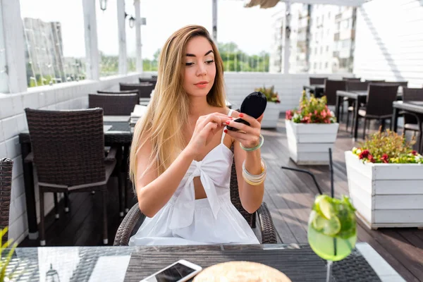 Beautiful and young girl in summer city cafe. In her hands she holds cosmetic bag with powder, paints herself with make-up. Business lady is preparing for meeting. Breakfast or snack at restaurant.