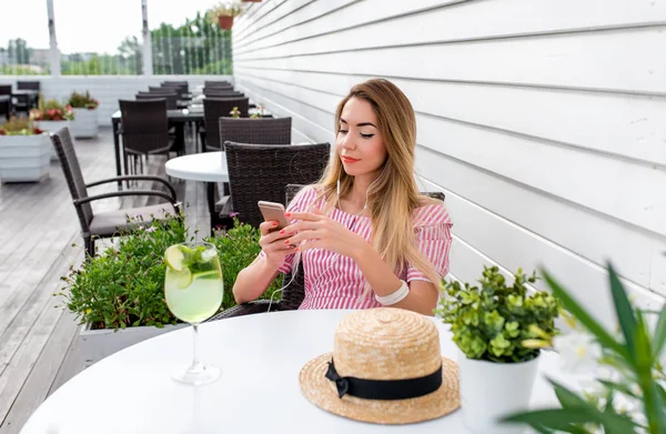 Girl in a dress sitting in a city cafe in summer. Calls a video call over the Internet. Happy enjoying music in headphones. Relax in the summer cafe on the veranda.