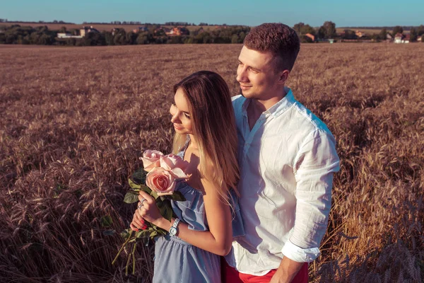 Feliz pareja joven hombre y mujer, familia romántica adulta. Conoce el atardecer en un campo de trigo. Feliz sonrisa. La chica en sus manos tiene un regalo, ramo de flores, de rosas . — Foto de Stock