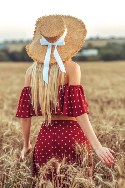 Flicka i en veteåker i sommaren i byn. Röd klänning och stråhatt, vy från baksidan. Kvinnan promenader på fältet. Begreppet gratis och fritid. — Stockfoto