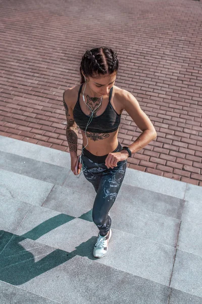 The girl athlete walks up stairs, in hands of a smartphone, listens to music in headphones. Looks at the clock, checks pulse and distance. Tanned skin and tattoos. The concept of a healthy lifestyle.