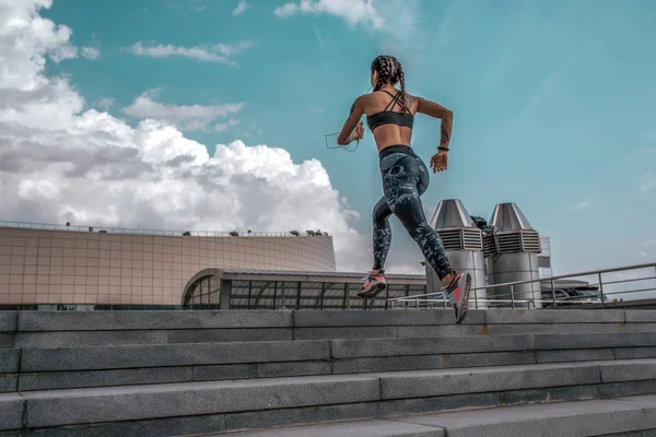 Спортсменка стрибає вгору по сходах, бігає на ранковий біг. Влітку в місті. Концепція здорового способу життя. Вид з тилу на тлі блакитного неба та бетонних сходів. Вільний простір для тексту . — стокове фото