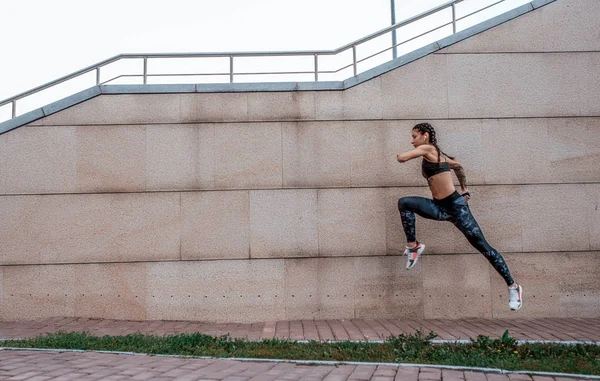 Дівчина-спортсмен стрибає, бігає на ранковий біг. Влітку в місті. Концепція здорового способу життя. Вид збоку на тлі бетонної стіни. Вільний простір для тексту . — стокове фото