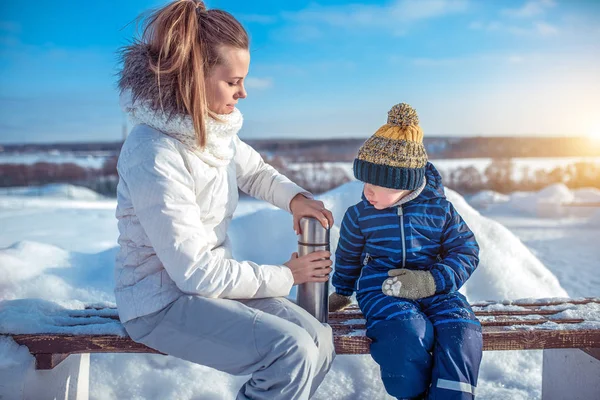 Молодая мать женщина открывает термос горячим супом или чаем, сидит на скамейке со своим сыном, мальчиком 2-5 лет. Отдых в зимнее утро на природе . — стоковое фото