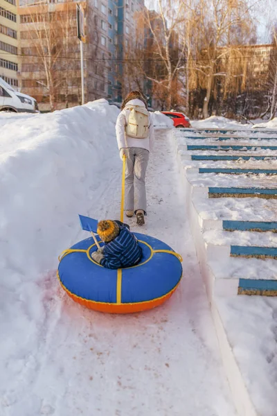 Woman in the winter in the city, drags a small child on a toy. Tubing on the belt. Rest on the hills in winter in the city.