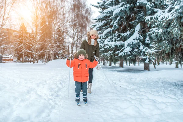 Langkah pertama dari seorang anak laki-laki bermain ski dengan tongkat. Happy tersenyum di taman saat musim dingin. Ibu pergi untuk seorang anak di belakang . — Stok Foto