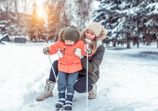 Langkah pertama dari seorang anak laki-laki bermain ski dengan tongkat. Bermain dan bersenang-senang di taman di musim dingin. Ibu terus mendukung anak, dan membantu untuk belajar berkuda dengan benar . — Stok Foto