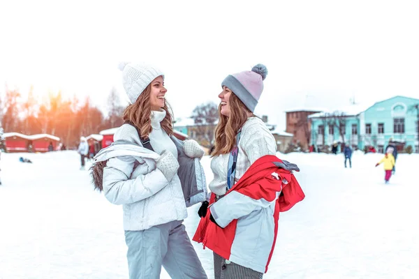Dwie piękne dziewczyny, rozmowy i żarty i mówienie, szczęśliwy, uśmiechnięty na zimowy dzień w parku miejskim. W zimowe ubrania i czapki na lodowisko. — Zdjęcie stockowe