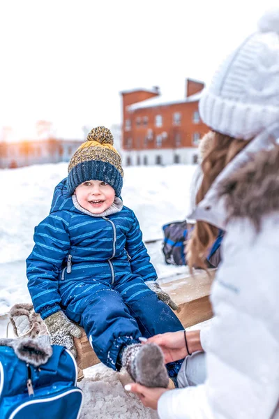 妈妈让儿子坐在冬季公园的长凳上。照顾一个年轻的母亲是一个孩子。穿袜子脱溜溜溜溜溜鞋. — 图库照片