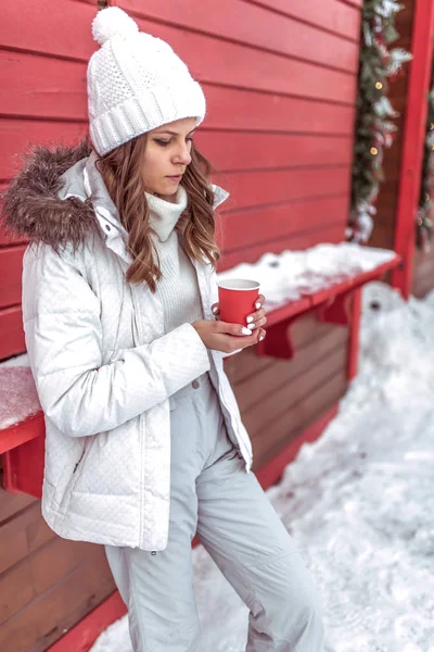 Piękna i młoda dziewczyna jest ocieplenie o kubku, trzymając kubek z gorącą kawę lub herbatę w jej ręce. W ciepły zimowy biały kapelusz i ubranie. Na tle czerwonego i pokryte śniegiem domów. — Zdjęcie stockowe