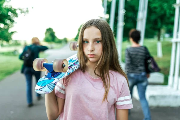 Adolescente en été en ville, triste debout dans le parc, bouleversée, tenant une planche à roulettes sur son épaule. De longs et beaux cheveux. Concept d'adolescents difficiles. Taches de rousseur sur le visage . — Photo