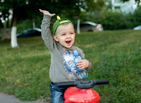Seorang anak kecil berusia 2-3 tahun, anak duduk di atas mainan. Di tangannya memegang botol air, teriakan bahagia. Di musim panas di taman kota . — Stok Foto
