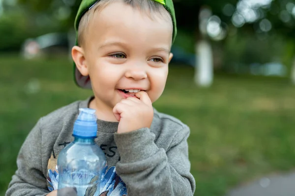 Um menino de 3-6 anos de idade, retrato close-up, a criança segura uma garrafa de água em sua mão, sorrindo feliz. No verão no parque da cidade . — Fotografia de Stock