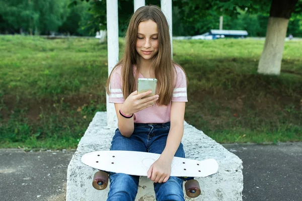 Seorang gadis remaja cantik berusia 10-12 tahun, duduk di taman kota di musim panas, memegang smartphone di tangannya, berkomunikasi di Internet melalui jaringan sosial. Rambut panjang di tangan skateboard . — Stok Foto