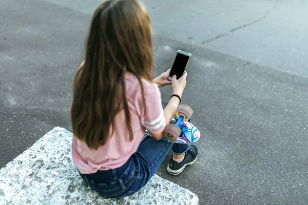 Девочка-подросток 9-11 лет, летом сидит в городском парке, держа в руках смартфон, общаясь по интернету через социальные сети. Длинные волосы вид сзади . — стоковое фото