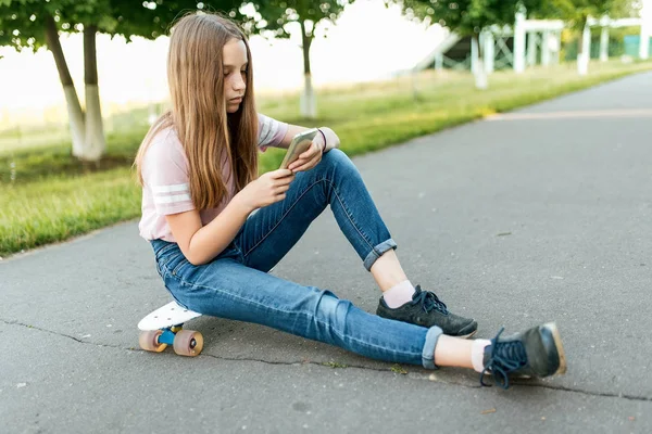 Gadis remaja berusia 8-13 tahun, duduk di skateboard di taman kota di musim panas, memegang smartphone di tangannya, berkomunikasi di jaringan sosial Internet. Rambut panjang, kecanduan internet . — Stok Foto