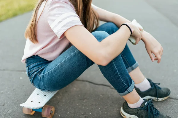 Gadis remaja berusia 7-14 tahun, duduk di skateboard di taman kota di musim panas, memegang smartphone di tangannya . — Stok Foto