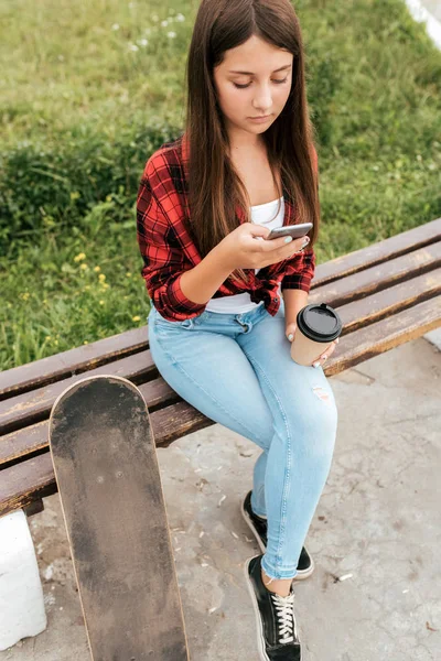 Sommer Teenager Mädchen sitzt auf einer Bank, hält ein Smartphone in der Hand und eine Tasse Kaffee. — Stockfoto