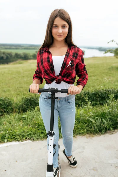 Gadis remaja dengan kemeja merah dan celana jeans, dengan skuter di musim panas di kota . — Stok Foto
