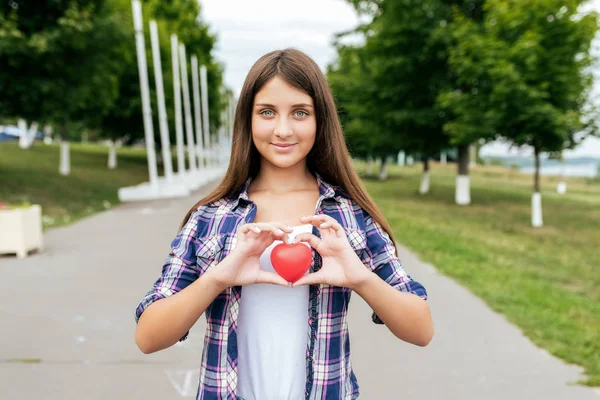 A teenager girl, 14-15 years old, stands in a city park in summer, holding a heart in her hands. In the summer, happy smiles. The concept of donation and transplant care for sick people.