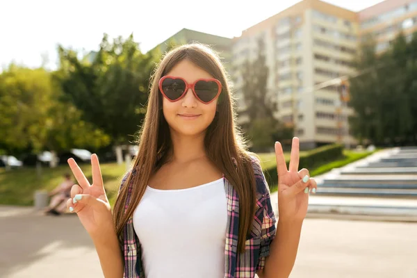 Tiener meisje 9-13 jaar oud, staan in de zomer in stadspark, hand gebaar Hallo overwinning. In de zomer, gelukkig glimlacht. Het concept van outdoor recreatie. In rood glazen in de vorm van een hart. — Stockfoto