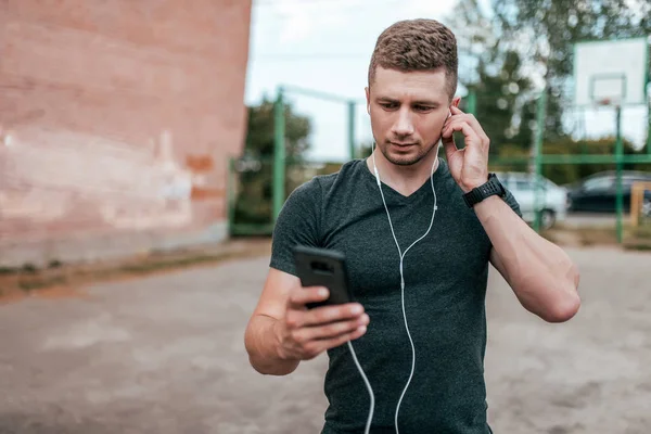 Молодой спортсмен, серьезный взгляд на экран смартфона, разговаривающий на видео. Лето в городском парке на фоне спортивной площадки. В повседневной одежде с наушниками, слушая музыку . — стоковое фото