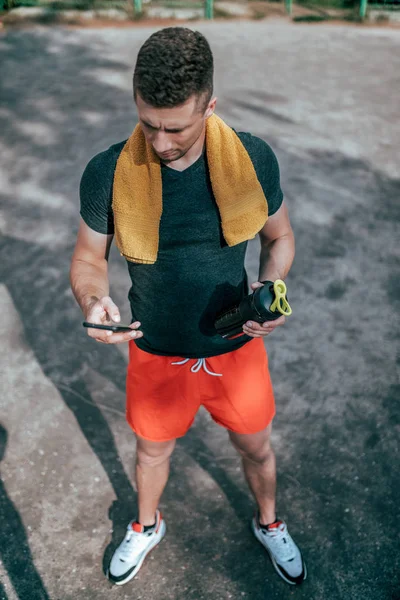 Ein junger Mann ist Sportler männlich, schaltet die Anwendung auf dem Smartphone-Bildschirm ein. Sommer im Stadtpark vor dem Hintergrund des Sportplatzes. in lässiger Kleidung mit Handtuch und Wasserflasche. — Stockfoto