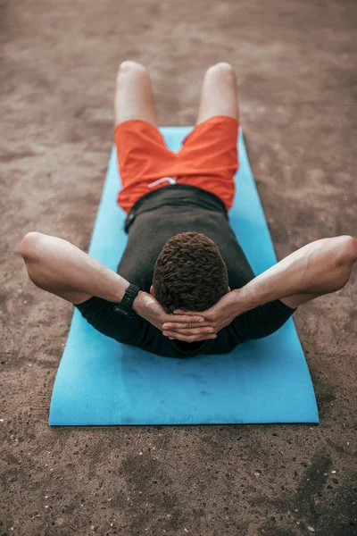 Male athlete, in the summer in city, lying on the athletic mat trains your abdominal muscles. Resting after a workout in the fresh air, in casual clothes and red shorts. Shakes the press.