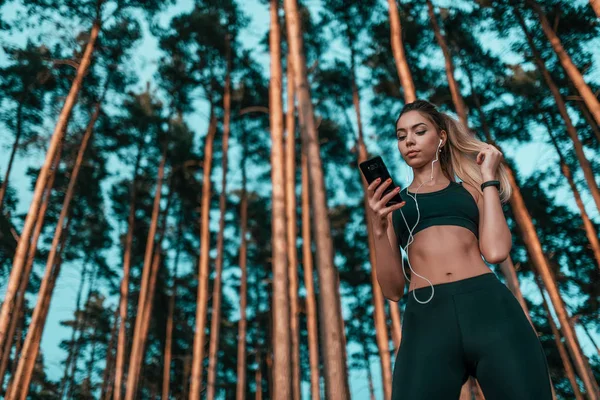 Gadis atlet cantik, berdiri di hutan latar belakang pohon, di udara segar. Gaya hidup aktif di taman musim panas. Pakaian olahraga kasual. Di tangan telepon dan mendengarkan musik di headphone. Ruang kosong untuk teks . — Stok Foto