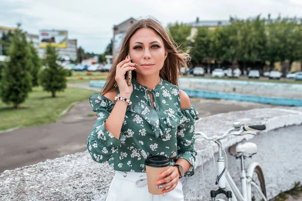 Mooi meisje brunette vrouw stond in het park achter de rug van de fiets. Telefoon in de hand een mok met warme koffie of thee. In de zomer in de stad tegen de achtergrond van de weg en groene bomen. — Stockfoto