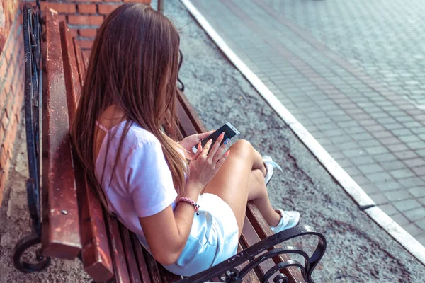 Mooi meisje brunette vrouw zitten op een bankje. Kijken naar video op de telefoon, wordt tekst afgedrukt. Zomer in de stad van de internettoepassing. — Stockfoto