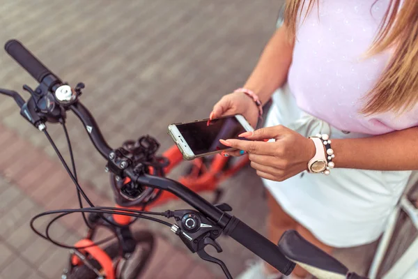 Mujer chica de pie en el parque en bicicleta. En verano, en la ciudad, en la mano de un smartphone, la aplicación desbloquea el aparcamiento de bicicletas junto a la aplicación. Primer plano. Anti-secuestro bloqueo bloqueador protector . —  Fotos de Stock