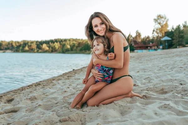 Krásná a šťastná mladá žena maminka objímání na břehu jezera s pískem, malé dítě dcera, šťastný úsměv, rodina odpočívá v letovisko na pobřeží. Pojem lásky a péče rodičů. — Stock fotografie