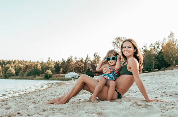 Krásná mladá žena a maminka, sedící na břehu jezera s pískem, v náručí malé dítě dceru, usmívající se, Rodina, odpočívá na letovisko na pláži. — Stock fotografie