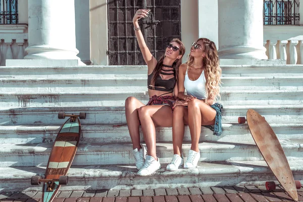 Zwei Mädchen im Badeanzug, Freundinnen, die im Sommer in der Stadt auf den Stufen sitzen. mit Longboard-Skateboard, Sonnenbrille. fotografiert Selfie-Handy-Anwendung auf einem Smartphone. Freiraum für Text. — Stockfoto