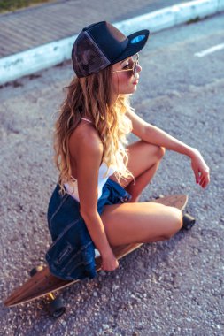 Bir beyzbol şapkalı bir kız oturuyor ve bir kaykay üzerinde longboard alır. Yazında şehre karşı bir asfalt yol, arkadan görüntüleyin. Uzun saçlı güneş gözlüğü.