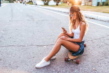 Bir beyzbol şapkalı bir kız bir kaykay üzerinde bir longboard oturur. Arka planda bir yol asfalt, Genç bir kadın şehirde yaz aylarında. Uzun saçlı güneş gözlüğü. Metin için boş alan.